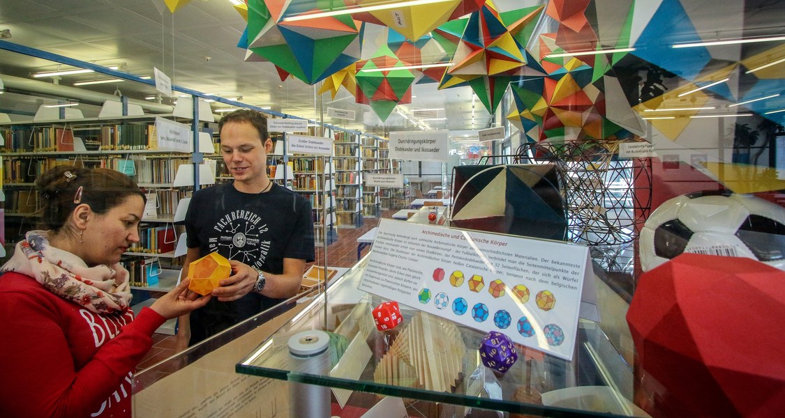 ein Student (rechts) und eine Studentin (links) schauen sich ein mathematisches Modell vor Regalreihen in der Bibliothek an
