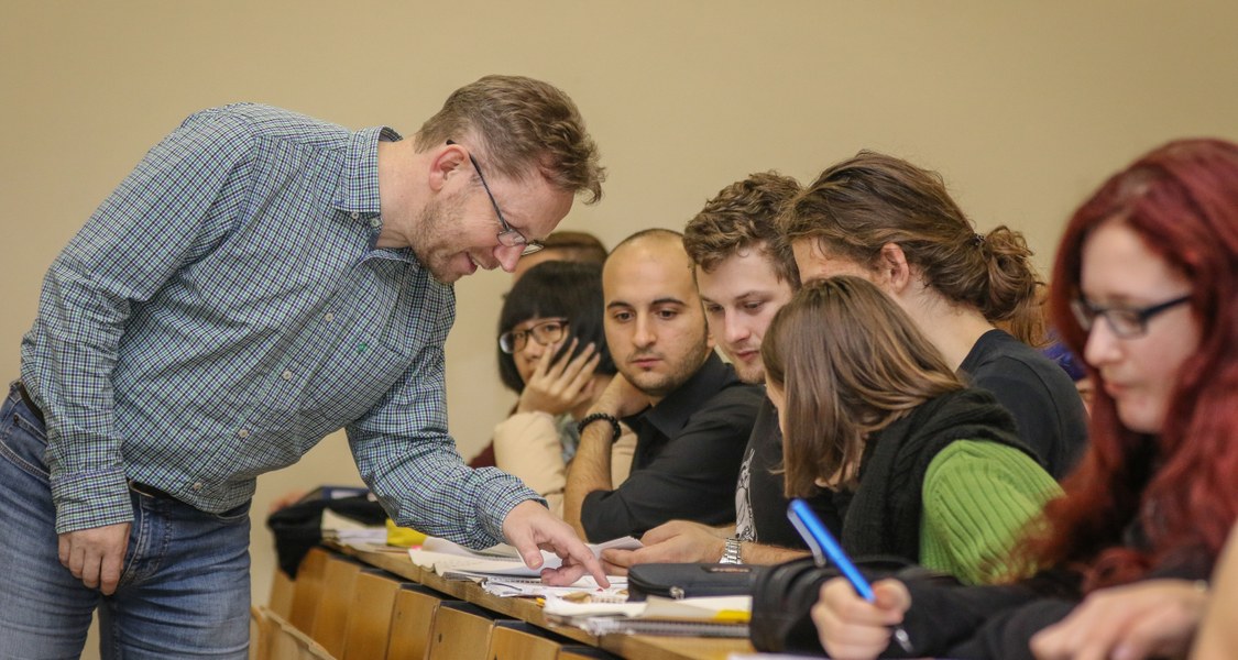 Studierende sitzen mit Schreibutensilien nebeneinander, vor ihnen erläutert ein stehender Dozent die Materie.