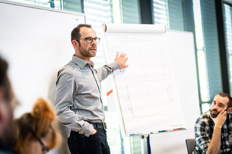 Dr. Reiner Becker steht vor einem Whiteboard und zeigt mit der linken Hand auf eine daneben stehende Pinnwand.