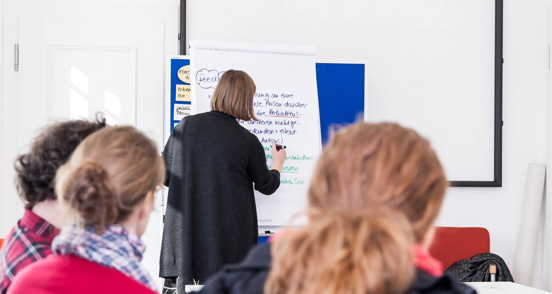 Menschen in einer Veranstaltung mit einer Dozentin, die an ein Whiteboard schreibt