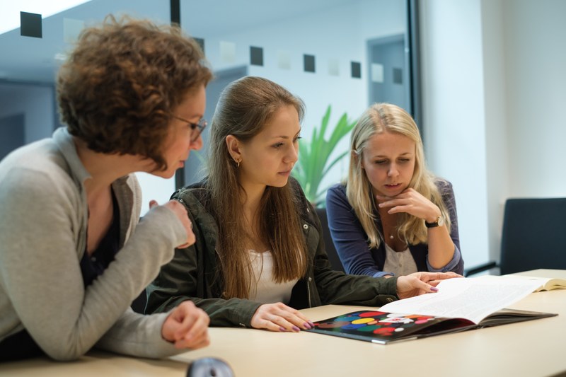 Drei Studentinnen sitzen in einem Seminarraum und schauen in ein Dokument.