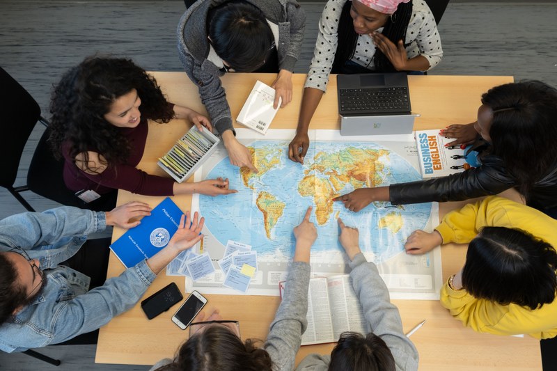 Studierende verschiedener Ethnien zeigen auf unterschiedliche Punkte einer Weltkarte in der Mitte des Tisches.