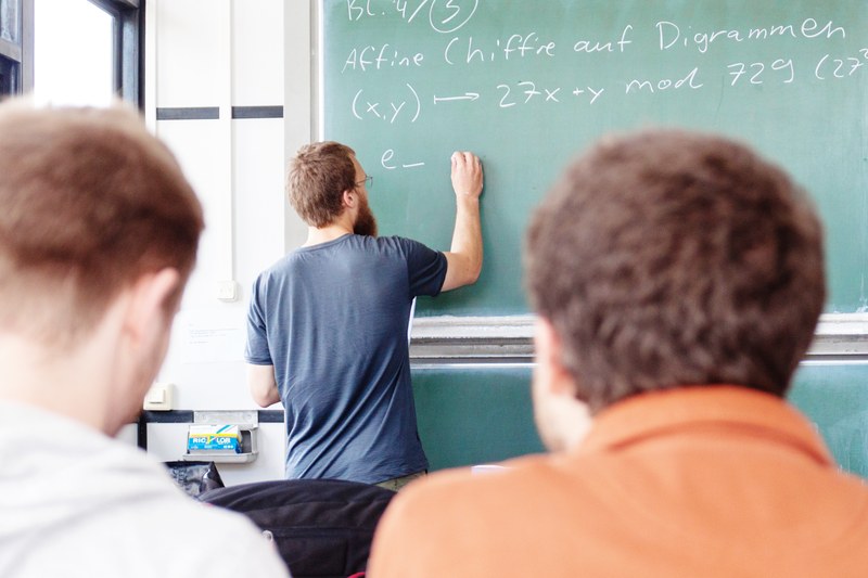 In einem Seminarraum schreibt ein Student mit Kreide mathematische Gleichungen an eine grüne Tafel.